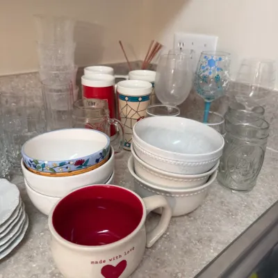 Sale Photo Thumbnail #66: Assorted kitchenware including ceramic bowls, coffee mugs, wine glasses, and glass tumblers arranged on a countertop.