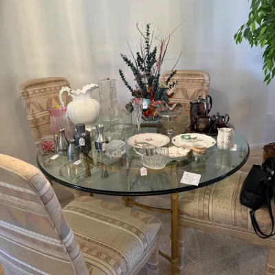 Sale Photo Thumbnail #62: Round glass-top dining table set with upholstered chairs, displaying an array of glassware, ceramics, silver-tone items, and decorative tableware.