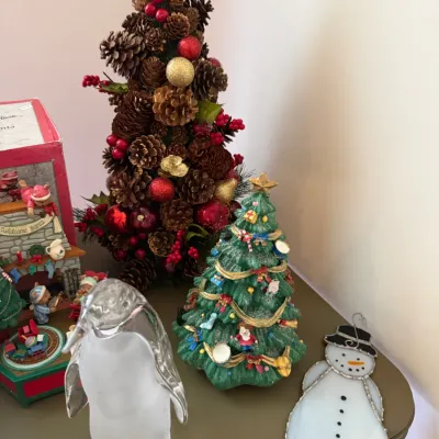 Sale Photo Thumbnail #25: Collection of holiday decor featuring a frosted glass penguin figurine, a detailed resin Christmas tree music box, a pinecone tree arrangement, and a stained glass snowman ornament.
