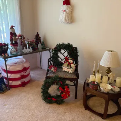 Sale Photo Thumbnail #60: Collection of Christmas decor including artificial wreaths, Santa Claus figures, decorative candles, a brass table lamp, and stacked storage containers displayed in a carpeted room.
