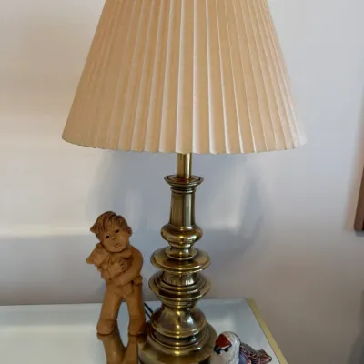 Sale Photo Thumbnail #10: Traditional brass table lamp with a pleated beige shade, displayed alongside a clay child figurine and a small bird figurine.