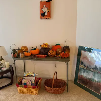 Sale Photo Thumbnail #59: Collection of seasonal fall and Halloween decor displayed on a folding table and end table, including artificial pumpkins, a wall hanging, woven baskets, candles, and framed landscape art.