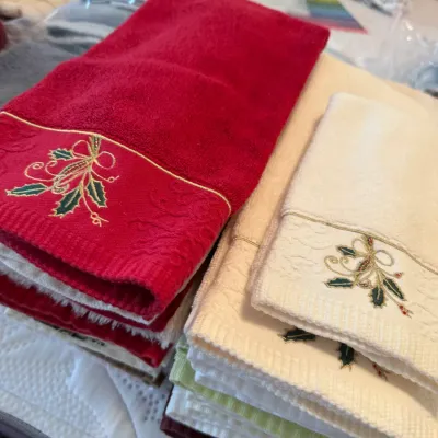 Sale Photo Thumbnail #55: Stacks of holiday-themed embroidered hand towels in red, cream, green, and white featuring holly leaf and gold ribbon motifs.