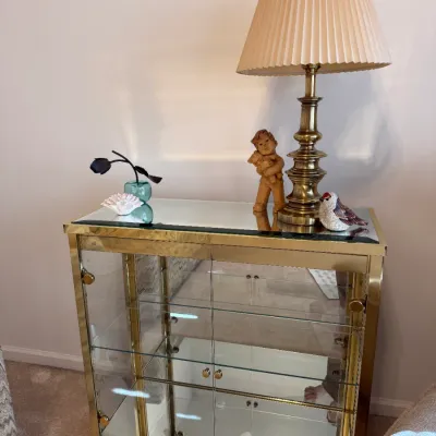Sale Photo Thumbnail #6: Vintage Hollywood Regency style brass and glass curio cabinet with mirrored back, topped with a brass table lamp with a pleated shade and small decorative figurines.