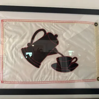 Framed nautical signal-style flag featuring a black coffee pot pouring into a teacup with red stitched outlines, presented in a black...