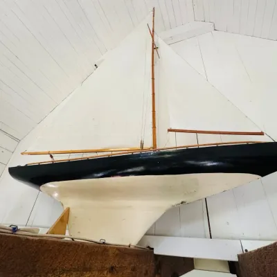 Large vintage wooden model sailboat featuring a black and cream painted hull, tall wooden spars, and intact white cloth sails mounted on...