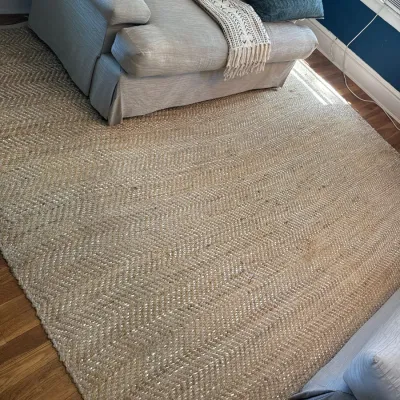 Large woven jute or sisal area rug with a herringbone pattern, displayed beneath neutral living room seating.