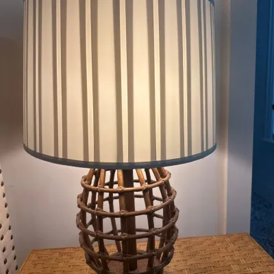 Boho-style table lamp with an open rattan cage base and a vertical striped drum shade, illuminated on a wicker surface.