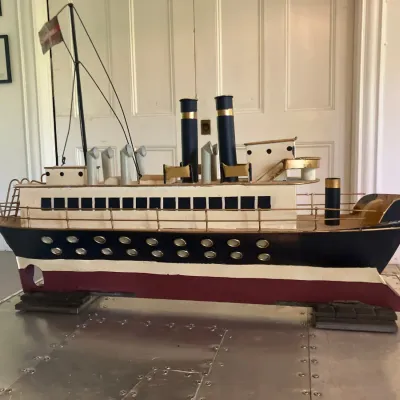 Vintage painted wooden model ocean liner featuring two smokestacks, brass railings, and small flags on a custom display stand.