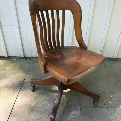 Vintage early-to-mid 20th-century wooden banker's desk chair featuring a curved slat back, contoured seat, and a four-star rolling...