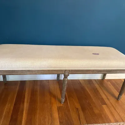 French Country style upholstered bench with weathered gray wooden fluted legs and neutral linen fabric seat.