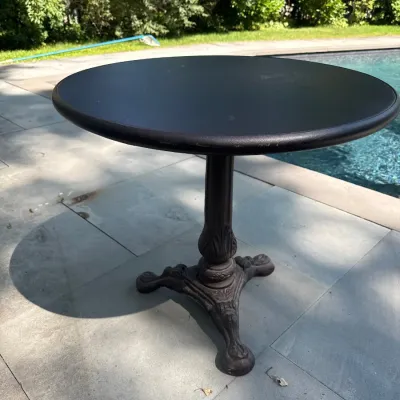 Black round bistro table with a smooth tabletop and an ornate, Victorian-style cast iron pedestal base.