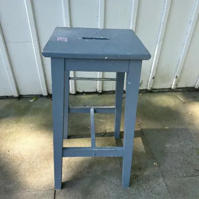 Simple grey-painted wooden stool with a center cutout handle on top, showing minor surface wear.