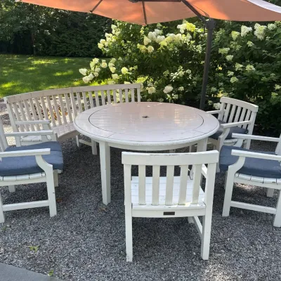 White wooden outdoor dining set featuring a round slat-top table, four armchairs with blue seat cushions, a curved bench, and a large...