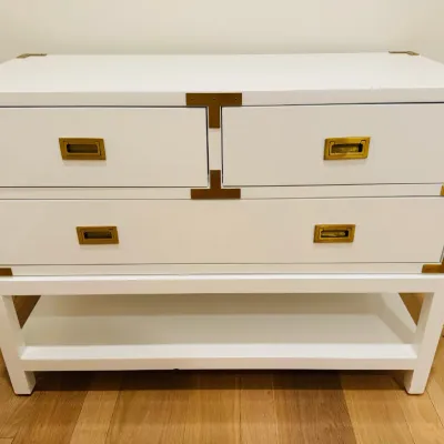 Sale Photo Thumbnail #16: White campaign-style chest of drawers with brass corner brackets and flush pulls, elevated on an open base with a lower shelf.