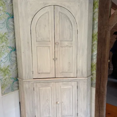 Sale Photo Thumbnail #32: Vintage white-washed wooden corner cabinet featuring arched upper doors, lower storage doors, and a detailed crown molding.