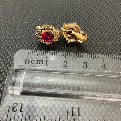Sale Photo Thumbnail #203: Pair of vintage gold-tone clip-on earrings featuring starburst or textured sunburst frames set with vibrant red/ruby-colored faceted round stones. Each earring measures approximately 1 cm wide according to the ruler shown. Good condition with light wear.