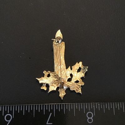 Sale Photo Thumbnail #116: A festive gold-tone Christmas brooch featuring a lit white candle with a red flame, surrounded by dark green enamel holly leaves and red berry accents. Standard pin back mechanism in good working order. Shows light vintage wear with intact enamel. Measure