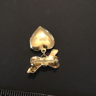 Sale Photo Thumbnail #101: Gold-tone vintage-style brooch featuring a ribbon bow motif with a dangling, engraved heart-shaped locket. The locket opens to hold small photos or keepsakes. Measures approximately 1.5 inches in total length. Features a secure pin-back clasp mechanism. L