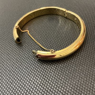 Sale Photo Thumbnail #142: Gold-tone polished hinged bangle bracelet featuring a classic rounded design, push-tongue clasp, and attached safety chain. Shows light surface wear and minor scuffs consistent with age and pre-owned use. Clasp and hinge function properly.