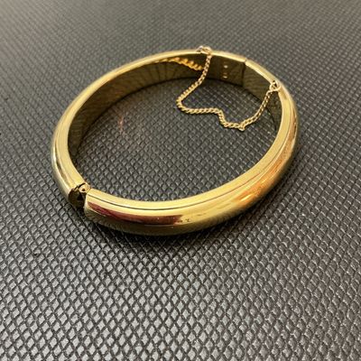 Sale Photo Thumbnail #139: Gold-tone polished hinged bangle bracelet featuring a classic rounded design, push-tongue clasp, and attached safety chain. Shows light surface wear and minor scuffs consistent with age and pre-owned use. Clasp and hinge function properly.