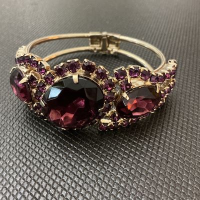Sale Photo Thumbnail #17: A statement vintage hinged bangle bracelet featuring deep purple/amethyst-colored prong-set rhinestones and large faceted center stones set in a gold-tone metal frame. Has a secure spring-hinge clasp. Measures approximately 2.5 inches wide. Overall in ver
