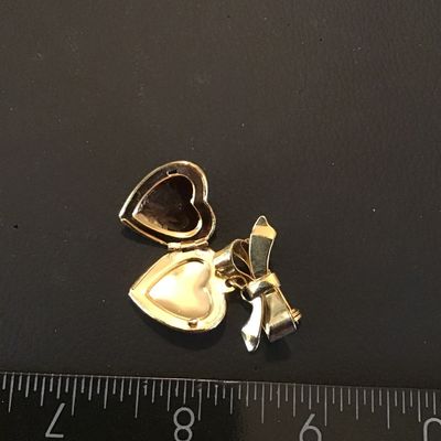 Sale Photo Thumbnail #98: Gold-tone vintage-style brooch featuring a ribbon bow motif with a dangling, engraved heart-shaped locket. The locket opens to hold small photos or keepsakes. Measures approximately 1.5 inches in total length. Features a secure pin-back clasp mechanism. L