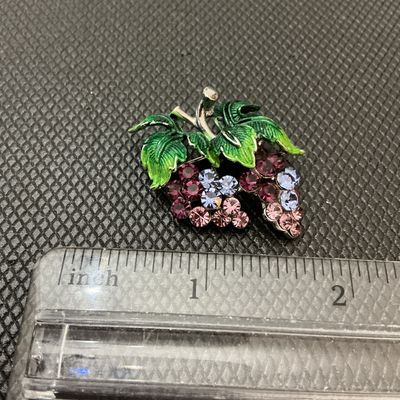 Sale Photo Thumbnail #9: A vintage costume jewelry brooch depicting a cluster of grapes. Features sparkling purple, lavender, and light pink rhinestones representing the fruit, topped with textured green enameled leaves and silver-tone stem detail. Measures approximately 1.5 inch