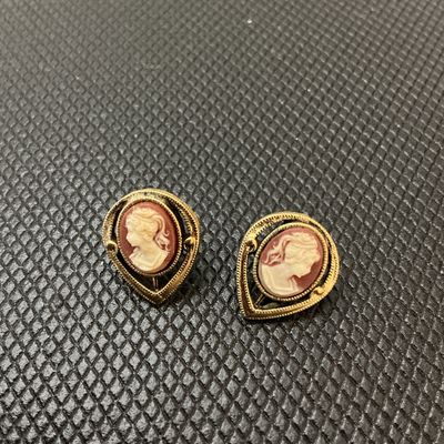 Sale Photo Thumbnail #188: Pair of vintage gold-tone pierced post earrings featuring a classic faux cameo portrait of a woman in profile set against a terracotta-colored background. The cameos are framed by detailed gold-tone openwork filigree teardrop settings. The backs display a