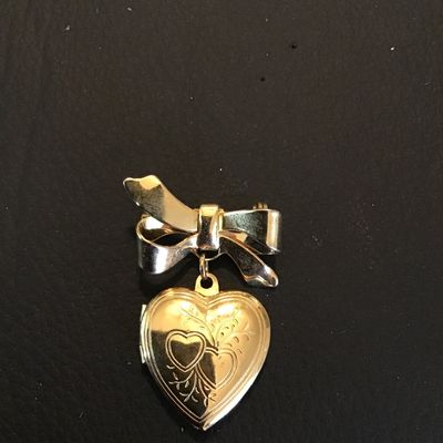 Sale Photo Thumbnail #95: Gold-tone vintage-style brooch featuring a ribbon bow motif with a dangling, engraved heart-shaped locket. The locket opens to hold small photos or keepsakes. Measures approximately 1.5 inches in total length. Features a secure pin-back clasp mechanism. L
