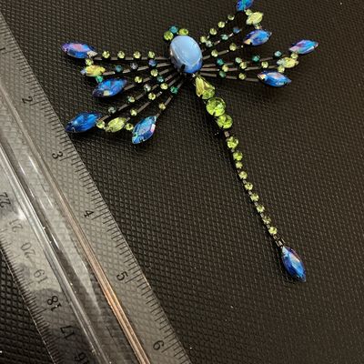 Sale Photo Thumbnail #2: A detailed statement dragonfly brooch set with vibrant blue, iridescent, and green/peridot rhinestones, featuring a light blue cabochon head. Set in dark gunmetal/black-toned metal with a pin back mechanism. Measures approximately 4.5 inches long by 3.5 i
