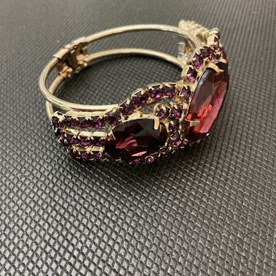 Sale Photo Thumbnail #16: A statement vintage hinged bangle bracelet featuring deep purple/amethyst-colored prong-set rhinestones and large faceted center stones set in a gold-tone metal frame. Has a secure spring-hinge clasp. Measures approximately 2.5 inches wide. Overall in ver