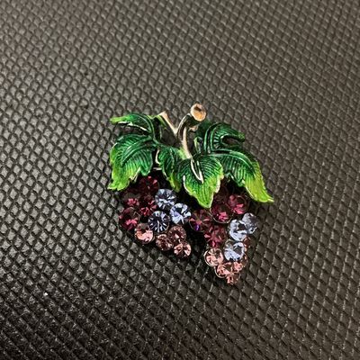 Sale Photo Thumbnail #8: A vintage costume jewelry brooch depicting a cluster of grapes. Features sparkling purple, lavender, and light pink rhinestones representing the fruit, topped with textured green enameled leaves and silver-tone stem detail. Measures approximately 1.5 inch