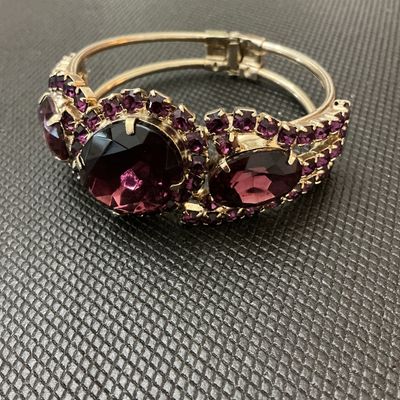 Sale Photo Thumbnail #12: A statement vintage hinged bangle bracelet featuring deep purple/amethyst-colored prong-set rhinestones and large faceted center stones set in a gold-tone metal frame. Has a secure spring-hinge clasp. Measures approximately 2.5 inches wide. Overall in ver