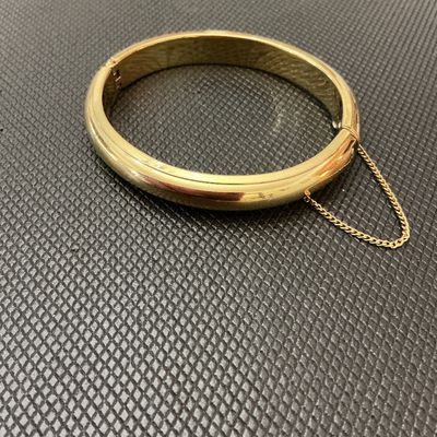 Sale Photo Thumbnail #136: Gold-tone polished hinged bangle bracelet featuring a classic rounded design, push-tongue clasp, and attached safety chain. Shows light surface wear and minor scuffs consistent with age and pre-owned use. Clasp and hinge function properly.