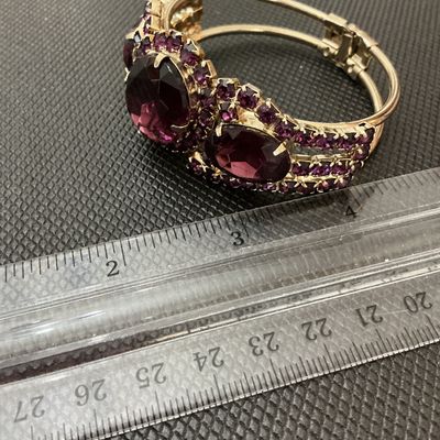 Sale Photo Thumbnail #19: A statement vintage hinged bangle bracelet featuring deep purple/amethyst-colored prong-set rhinestones and large faceted center stones set in a gold-tone metal frame. Has a secure spring-hinge clasp. Measures approximately 2.5 inches wide. Overall in ver