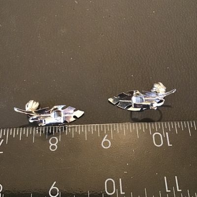 Sale Photo Thumbnail #171: Pair of vintage silver-tone clip-on earrings featuring a textured feather or leaf design with a small detailed flower or acorn accent. Each earring measures approximately 1.5 inches in length. In good vintage condition with working clip backs.