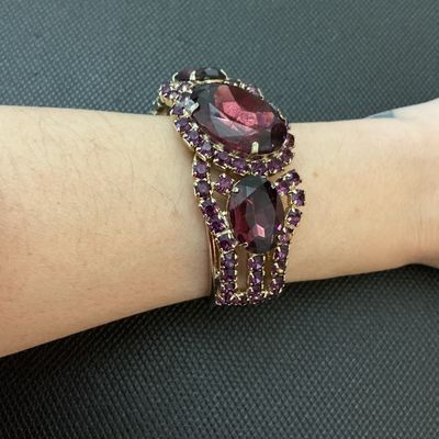 Sale Photo Thumbnail #14: A statement vintage hinged bangle bracelet featuring deep purple/amethyst-colored prong-set rhinestones and large faceted center stones set in a gold-tone metal frame. Has a secure spring-hinge clasp. Measures approximately 2.5 inches wide. Overall in ver