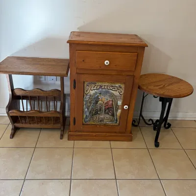 Sale Photo Thumbnail #51: Trio of wooden side tables, including a pine cabinet with a painted 'Farmers Backbone of America' graphic door panel, flanked by a wooden magazine rack table and an oval top table on a black wrought iron base.