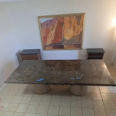 Sale Photo Thumbnail #10: Large dark brown marble dining table with thick pedestal bases, featured alongside a framed desert landscape print and small side cabinets.