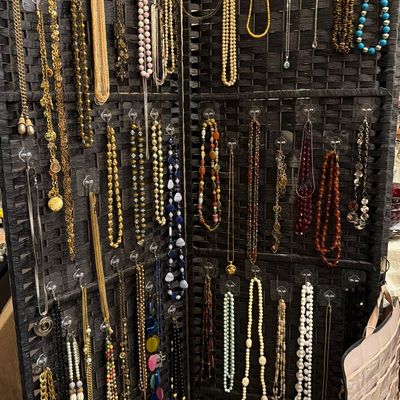 Large collection of vintage costume jewelry necklaces arranged on a dark woven folding screen, featuring beaded, gold-tone chain, and...