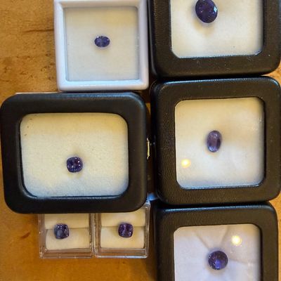 Collection of loose, faceted violet-blue gemstones, likely tanzanite or sapphire, presented in individual display cases.