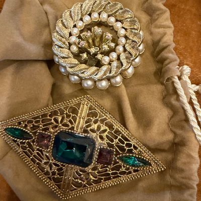 Pair of vintage costume brooches resting on a drawstring pouch, including a textured gold-tone wreath brooch with faux pearls and a...