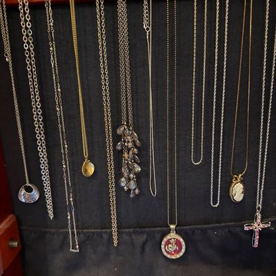 Collection of assortment pendant necklaces hanging in a wooden jewelry cabinet, including a cameo pendant, cross necklace, and crystal...