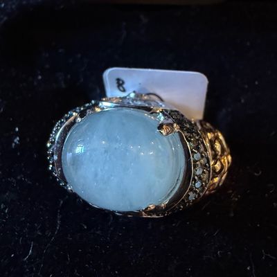 Sale Photo Thumbnail #22: Statement ring featuring a large oval pale blue cabochon stone set in a detailed, stone-encrusted metallic band.