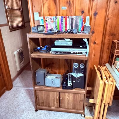 Sale Photo Thumbnail #75: Wooden media shelving unit, various electronics, VCR and DVD players, computer speakers, power strips, and a collection of VHS tapes and books.