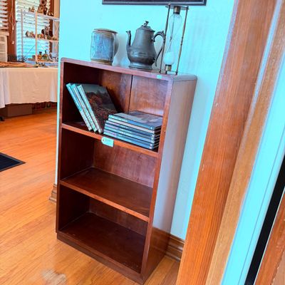 Sale Photo Thumbnail #24: Vintage wood four-shelf bookcase holding assorted hardbound books, topped with a pottery crock, metallic teapot, and brass sand timer.