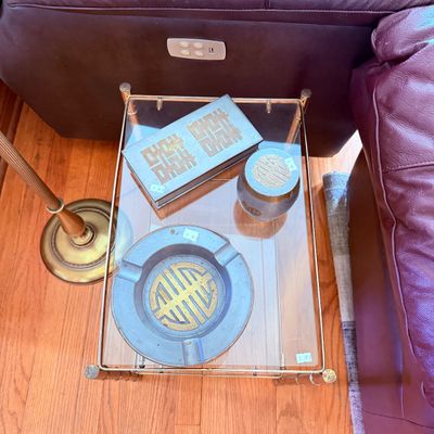 Sale Photo Thumbnail #23: Pewter smoking accessory set with brass Chinese character motifs, including a rectangular cigarette box, lidded jar, and ashtray, displayed on a glass-top side table.