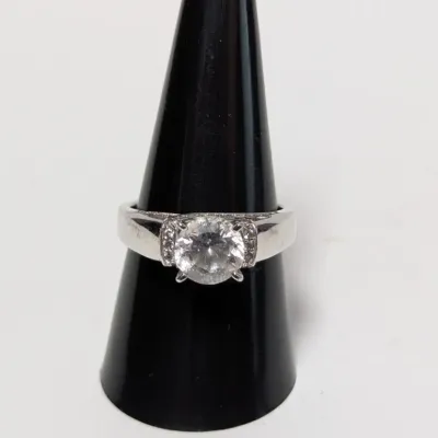 Sale Photo Thumbnail #952: Solitaire-style ring featuring a round-cut clear stone set in a wide silver-tone band with small accent stones on the shoulders, displayed on a black ring cone.