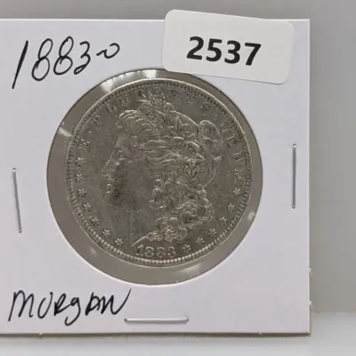 Sale Photo Thumbnail #1028: 1883-O Morgan silver dollar coin, housed in a cardboard flip holder with handwritten and printed labels.